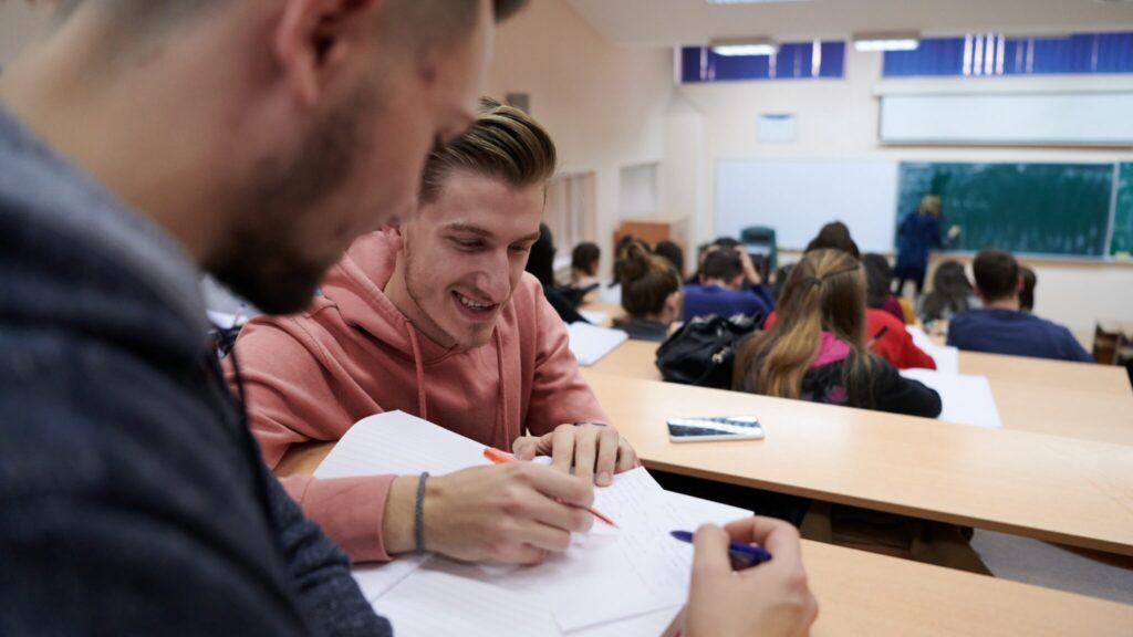 Two male students collaborate on notes in a lecture hall while others listen to the instructor at the front of the room.