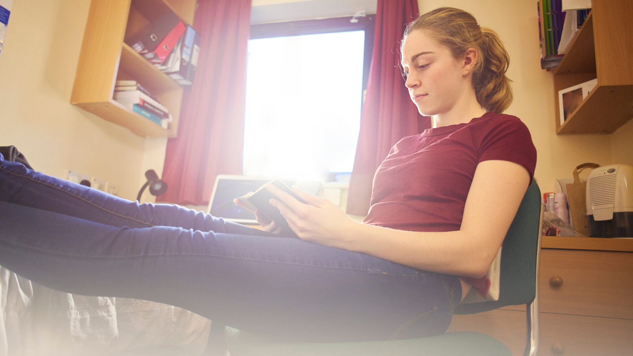 Girl in university bedroom sat with feet up and book in hand