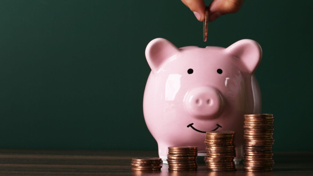Pink pig money box with hand putting coin in and a pile of coins in front of the pig