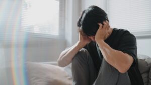Young person in black t-shirt and grey joggers sat on a bed with their head in their hands.