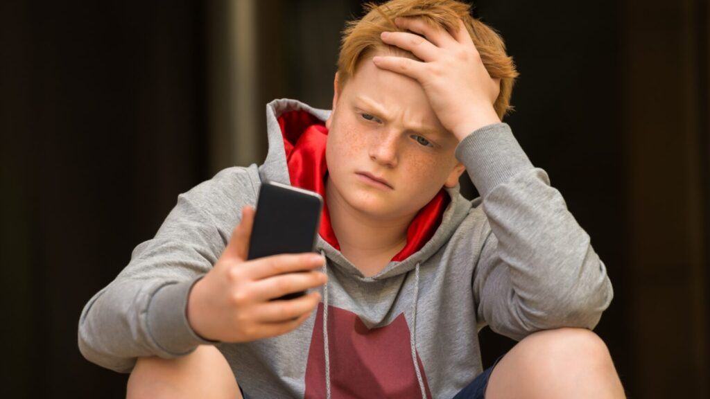 Teenage boy sat on floor, holding phone in one hand and his head in another