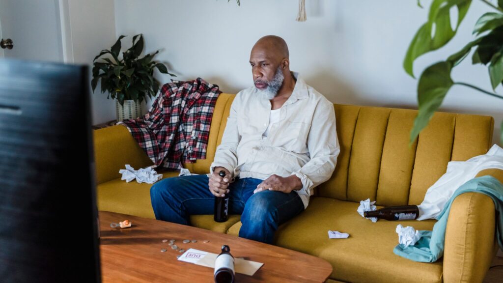 Man sat alone, looking sat on a sofa in the living room. Her is surrounded by alcohol - empty bottles of beer fill the table and sofa next to him. The man stares dazed into the TV.
