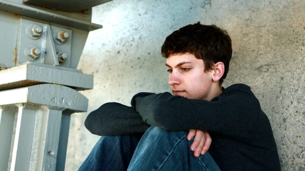 Teen boy slumped sat against a wall with his arms hugging his knees