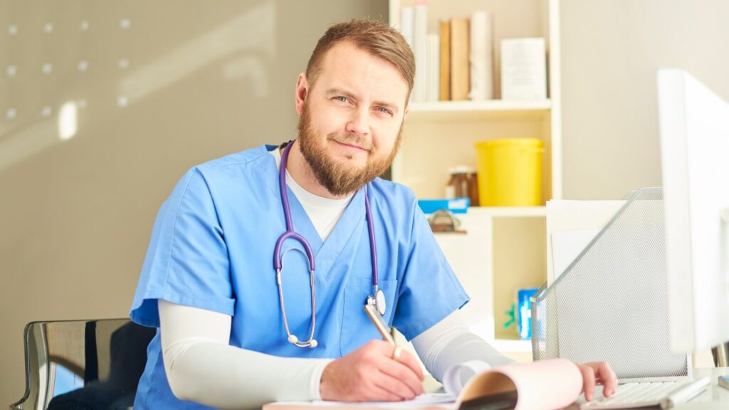 Doctor wearing blue scrubs smiling whilst writing notes