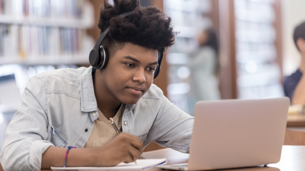 Older teenage boy working at a desk wearing a pair of headphones. His laptop is open.