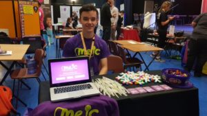 A Meic stand set up at an event with a laptop on the table. A person is sat behind the table in a Meic branded purple tshirt